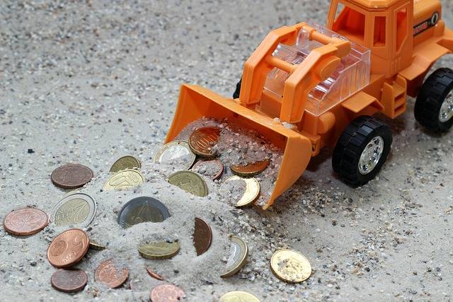 Ein oranger Spielzeugbagger schiebt einen Zuschuss zur Klassenfahrt in Münzen im Osteesand zusammen.